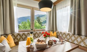 A cozy dining area with a wooden table and sunny view. On the table are fresh rolls, fruit, and drinks.