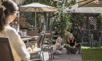 A relaxed garden scene with a woman sitting and two girls playing with a cat. In the background, umbrellas and flowering plants are visible.