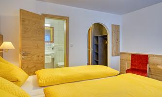 A cozy room with two beds and yellow bedding. In the background, a bathroom is visible, and there is a corner with a red chair.