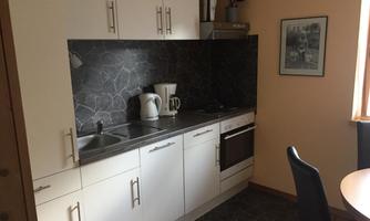 A small modern kitchen with white cabinets and a gray tile backsplash. On the countertop, there is a kettle and various kitchen utensils.