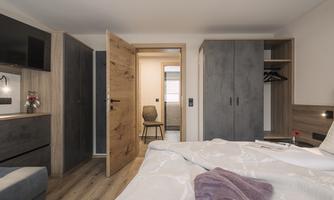 A modern bedroom with a large bed and wood paneling. In the background, a wardrobe and a chair are visible, as well as an open room with a door.