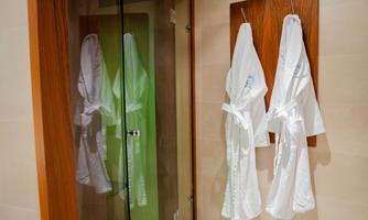 Two white bathrobes hang on a wooden panel. In the background, there is a glass shower cabin.