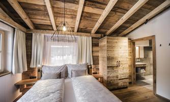 A cozy bedroom with a wooden ceiling and natural light. The decor is modern and rustic, with a view of an adjacent bathroom.