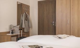 A cozy hotel room with two beds and a wooden wardrobe. In the background, there is a mirror as well as a coat rack with a coat.