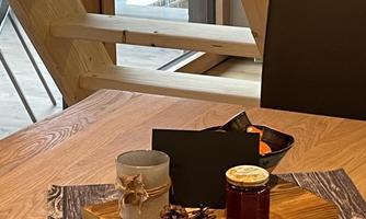 A stylish table with a wooden board on which a glass of honey, a candle, and some pine cones are placed. In the background, wooden structures and windows can be seen.