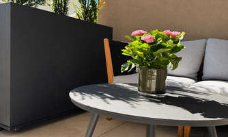 A modern terrace with a round table and a cozy sofa. On the table, there is a plant. Behind it, you can see the privacy screen for sufficient privacy.
