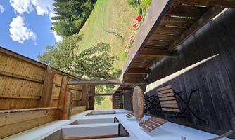 A cozy balcony with wooden chairs and a small table. In the background, there is a green meadow and trees under a blue sky.