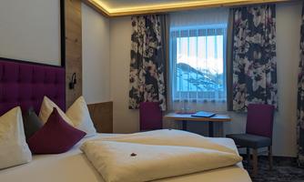 A cozy hotel room with a large bed and colorful curtains. Through the window, you can see the mountains in the background.
