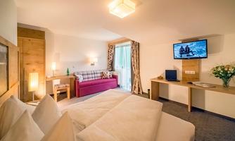 A cozy room with two beds, a sofa bed, and a television. Large windows provide plenty of daylight and a pleasant atmosphere.