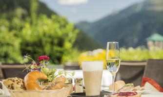 A set table with fresh rolls, a cup of coffee, and a glass of sparkling wine. In the background, green mountains and a blue sky can be seen.