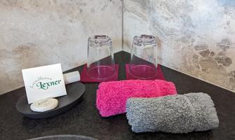 A modern-designed room with two glasses, a pink towel and a gray towel, as well as a soap on a small plate. A note about a wellness service is also present.