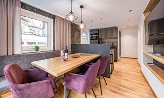A modern dining room with a wooden table and purple chairs. The walls are bright, and the windows let in plenty of light.
