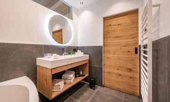 A modern bathroom with a wooden drawer, a sink, and a round mirror. The walls are painted in neutral colors, and the door is made of wood.