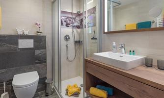 A modern bathroom with a shower, sink, and wooden shelf. Towels and toiletries are neatly arranged.