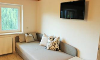 A cozy living room with a gray sofa and decorative cushions. A television hangs on the wall, and through the window, there is a view of the green trees outside.