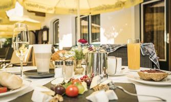 A laid table with a selection of breakfast dishes, including fresh fruit, cheese, and rolls. There is also champagne and orange juice, surrounded by sunny umbrella spots.