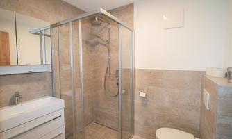 A modern bathroom with a glass shower cabin. The walls are designed with beige tiles, and there is a wash area with a large mirror.
