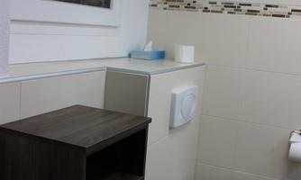 A modern bathroom with a WC, a wooden shelf, and a small window opening. The walls are tiled in light colors and decorated with a narrow strip of black and gray tiles.