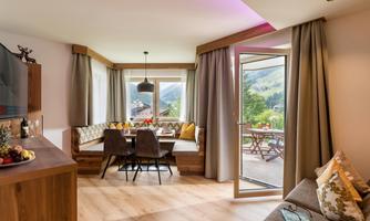 A cozy living area with a dining group and large windows overlooking a green landscape. Bright curtains and warm wood give the room an inviting atmosphere.