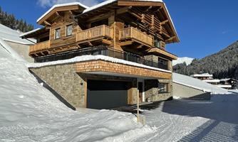 A cozy wooden house with large balconies, surrounded by snow-covered slopes. The clear blue sky completes the winter landscape.