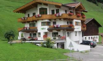 A traditional Alpine house with balconies and blooming flowers. It stands in a green landscape with a clear sky.