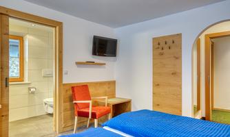 A modern bedroom with blue bedcovers and a television on the wall. In the background there is a bathroom and a small table with a chair.