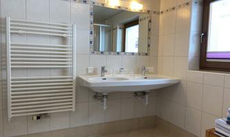 A modern bathroom with a double sink and a large mirror. On the wall, there is a towel radiator and a window provides natural light.