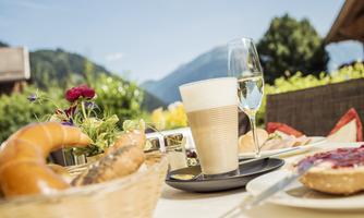 A beautiful breakfast table outdoors with fresh croissants, a cup of coffee, and a glass of champagne. In the background, you can see green mountains and blooming plants.