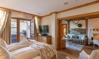An elegant bedroom with two beds and wooden furniture. In the background, there is a cozy living area with a seating area and a balcony.