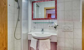 A modern bathroom with a shower, sink, and mirror. The walls are tiled and there are towels on the shelf.