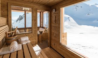 A modern sauna with wooden interior and large window view of snow-covered mountains. White towels and a small cooler are provided, ensuring relaxation.