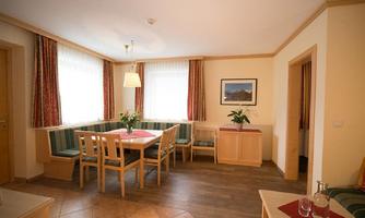 A bright and inviting dining room with a large table and wooden chairs. The walls are adorned with curtains and a picture, and fresh flowers are on the table.