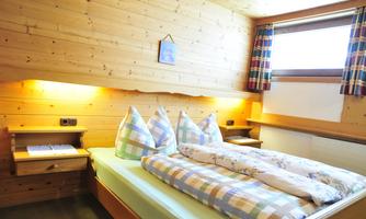 A cozy bedroom with wood paneling and two beds. The windows are fitted with checked curtains and soft light brightens the room.