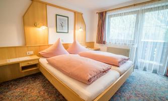 A cozy hotel room with two single beds and pink pillows. Large windows let in plenty of light and offer a view of the outdoors.