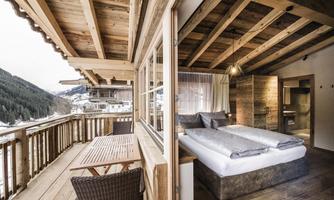 A cozy room with wooden furnishings and a large window. The balcony offers a view of the mountains.