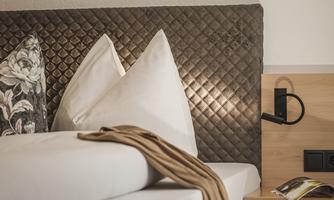 A cozy bedroom with a modern headboard and white pillows. Next to the bed is a reading lamp and a small shelf.