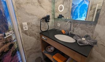 A modern bathroom with a sink on a wooden dresser. The walls are decorated with gray tiles, and there is a mirror with a lit frame.