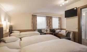 A bright room with three beds and a table. Large windows provide plenty of daylight.