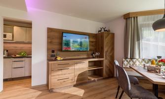 A modern living room with wooden furniture and a television on the wall. The room has large windows and offers a bright, inviting atmosphere.