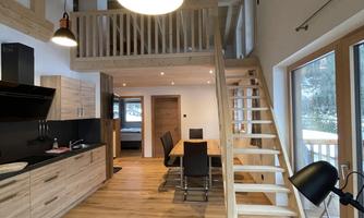 A modern, bright living room with wood details and an open staircase. The large windows provide a beautiful view outside.