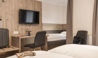 A bright hotel room with wooden paneling, a television, and a desk. There are two beds and a suitcase in the corner.