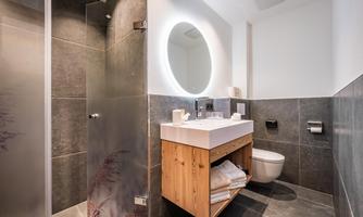 A modern bathroom with gray tiles and a glass shower. In the foreground is a wooden sink with a large, round mirror.