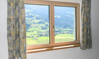 A window with a wooden frame and curtains, showcasing a picturesque landscape. Green meadows and gentle hills are visible in the background.