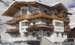 A modern mountain chalet with timber cladding and multiple balconies. It is situated in a snowy landscape, surrounded by mountains.