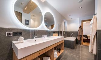 A modern bathroom with a double sink and large illuminated mirrors. The space is elegantly designed with wood and stone accents.