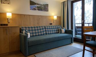 A cozy living room with a blue sofa and a dining table. Large windows provide plenty of daylight and a view outside.