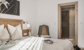 A modern bedroom with a cozy bed and bright bedding. In the background, there is a bathroom with a glass shower cubicle.