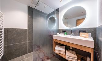 A modern bathroom with a spacious shower and a round mirror. The wooden table offers fresh towels and bathroom items.