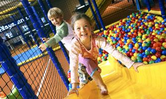 A cheerful child slides down the slide, while another child plays in the background. Colorful balls are scattered in one area of the playground.