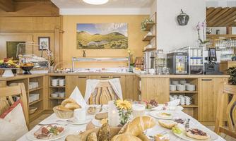 A cozy breakfast room with wooden interiors. On the table, there is an assortment of rolls, cold cuts, cheese, and fresh fruits.